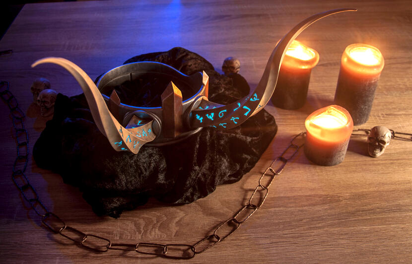 Photo du props "couronne de roi démon". La couronne est posée sur un tissu noir sur une table en bois, des bougies sont à côtés. Une chaîne traverse la table. La couronne a deux cornes sur lesquelles des runes lumineuses bleues sont gravées.